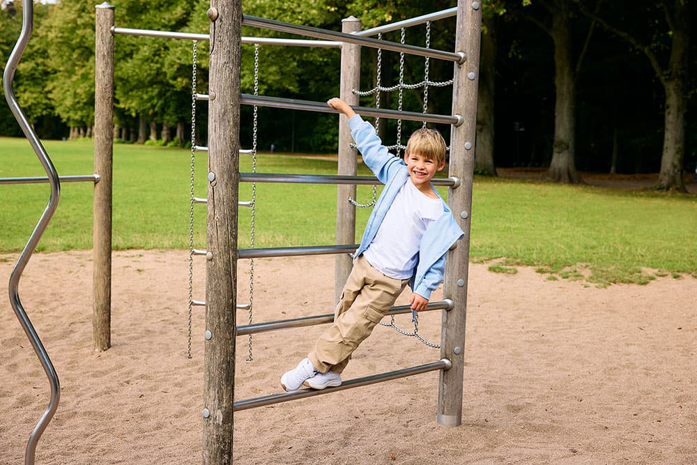 Ein Kind in einer hellblauen Jacke klettert lächelnd auf einem Klettergerüst, im Hintergrund sind Bäume und Gras zu sehen.