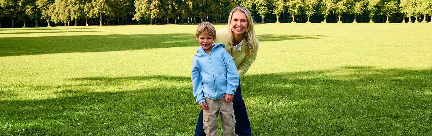 Eine Frau und ein Kind in einer blauen Jacke stehen auf einer Wiese mit Bäumen im Hintergrund und lächeln in die Kamera.