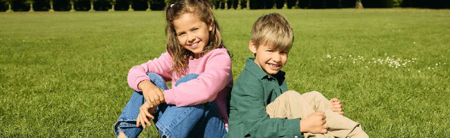 Two children, a girl and a boy, sit on grass in a sunny park. She wears a pink jacket, and he wears a green one. Both smile at the camera.