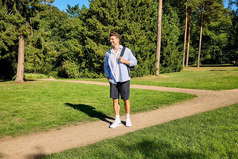 Ein Mann in einem blauen Hemd und Shorts geht auf einem Kiesweg durch einen grünen Park, umgeben von Bäumen und Gras.