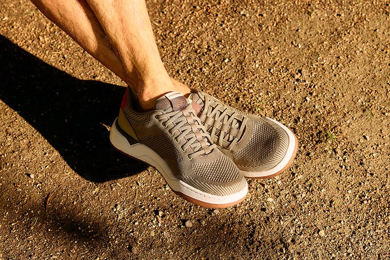 Eine Person in beigen Mesh-Sneakers steht auf einem sonnenbeschienenen Feldweg und wirft einen Schatten nach links.
