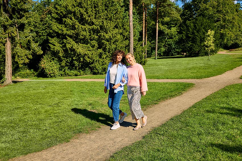 Zwei Frauen spazieren auf einem Weg in einem Park, umgeben von grünen Bäumen und Wiesen unter einem strahlend blauen Himmel.
