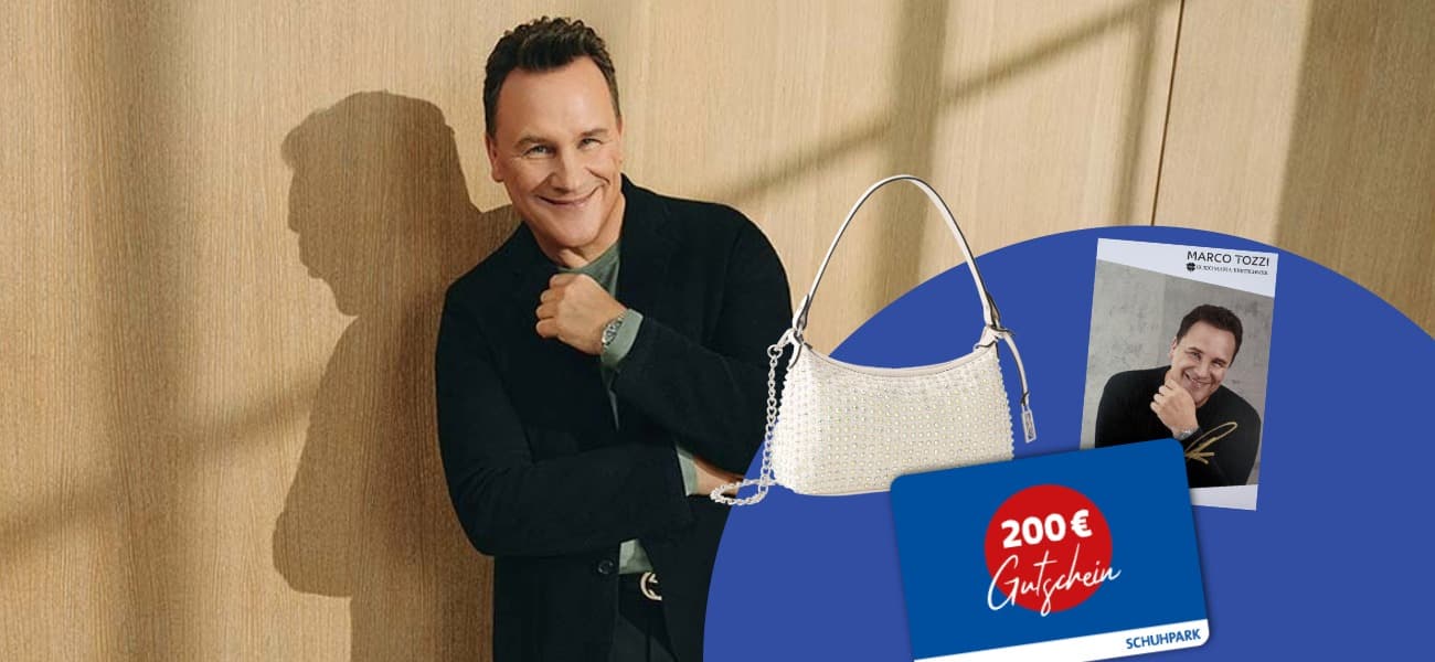 Ein lächelnder Mann (Guido Maria Kretschmer) in dunkler Kleidung, eine Handtasche, ein signiertes Foto und ein 200-Euro-Gutschein vor einem holzfarbenen und grauen Hintergrund.
