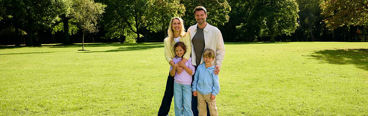 Eine vierköpfige Familie steht lächelnd auf einem grünen Rasen, umgeben von Bäumen in einem sonnigen Park.