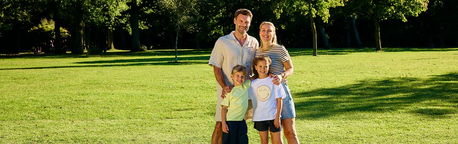 Eine lächelnde vierköpfige Familie steht zusammen auf einer sonnigen Wiese mit Bäumen im Hintergrund.