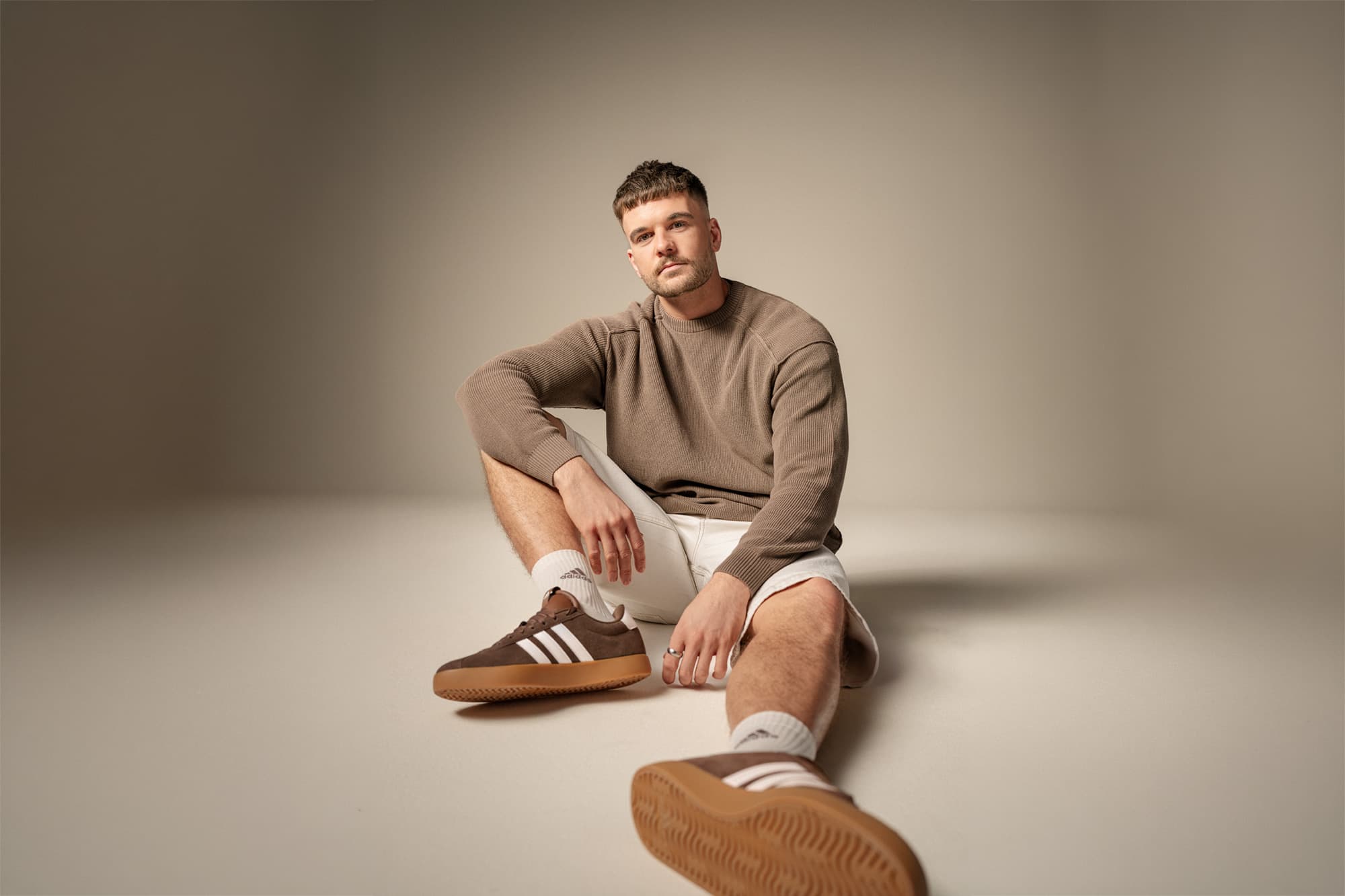 Mann sitzt auf dem Boden und trägt einen braunen Pullover, weiße Shorts und braune Turnschuhe vor einem neutralen Hintergrund.